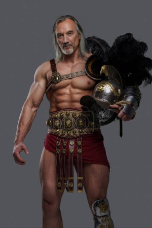 Téléchargez les photos : Vieilli mais toujours puissant, ce gladiateur musculaire aux longs cheveux gris et à la barbe se dresse fièrement en armure légère sur fond gris - en image libre de droit