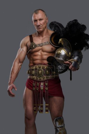 Photo for Muscular gladiator of mature age with a wrinkled face wears lightweight yet ornate armor, holding a gladiator helmet with a feathered crest as he poses confidently against a grey background - Royalty Free Image