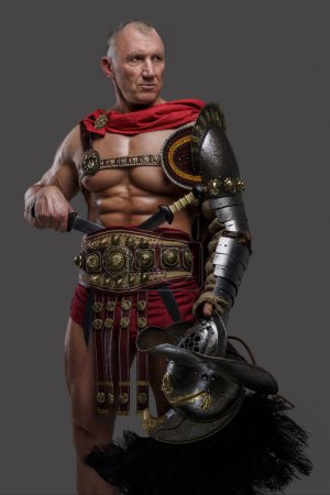 Photo for Experienced and muscular gladiator of mature age with wrinkled face wears lightweight armor, holding a gladiator helmet with a feathered crest as he poses confidently against grey background - Royalty Free Image