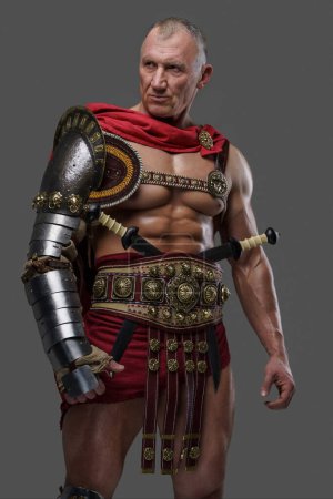 Foto de Gladiador experimentado y musculoso de años maduros con una apariencia áspera lleva armadura mientras se levanta con confianza sobre un fondo gris - Imagen libre de derechos