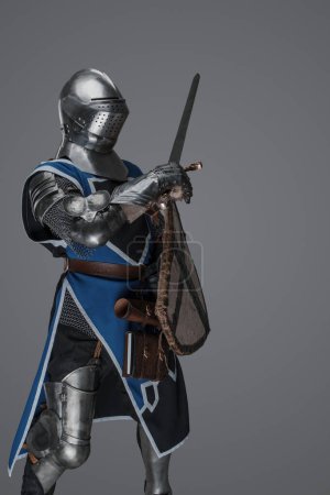 Foto de Caballero medieval en pose de batalla activa, empuñando espada y escudo contra fondo gris - Imagen libre de derechos