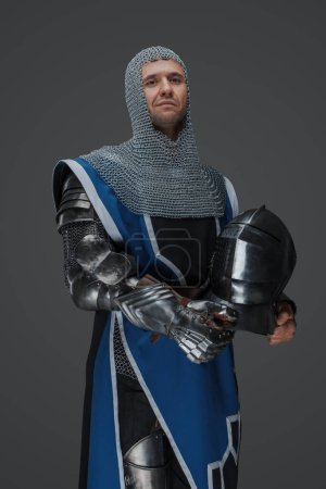 Photo for Royal guard dressed in medieval armor and blue surcoat, with chain mail coif, standing holding helmet in hand, against a gray background - Royalty Free Image