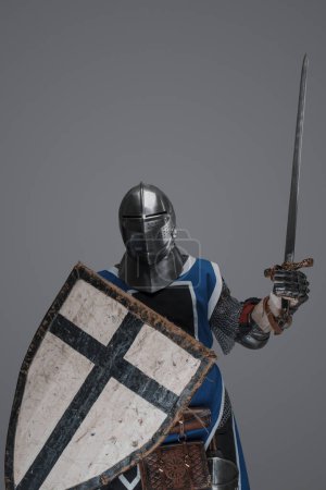 Photo for Medieval knight in active battle pose, wielding sword and shield against gray background - Royalty Free Image