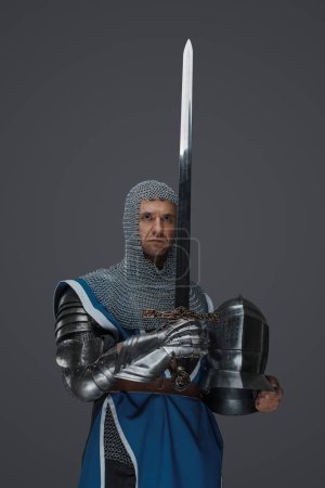 Téléchargez les photos : Fier chevalier tient son épée et son casque, revêtu d'un survêtement bleu, d'une coiffe en chaine et d'une armure médiévale, sur fond gris - en image libre de droit
