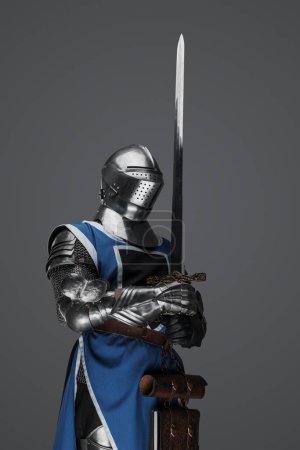 Foto de Guardia medieval vestida con armadura y sobretodo azul sosteniendo una espada, de pie en pose de estatua, sobre un fondo gris - Imagen libre de derechos