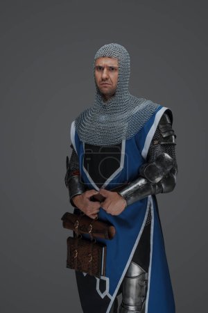 Téléchargez les photos : Guerrier médiéval vêtu d'une armure brillante, d'un survêtement bleu et d'une capuche en chaine, tenant une épée, debout résolument sur un fond gris neutre - en image libre de droit