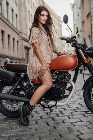 Foto de Impresionante chica de ojos azules se sienta en su motocicleta de estilo retro en una vieja calle empedrada en Europa - Imagen libre de derechos