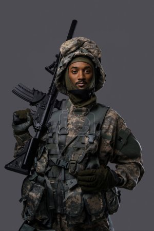 Photo for Dark skinned soldier in NATO uniform and helmet poses with a serious expression on a plain grey background - Royalty Free Image