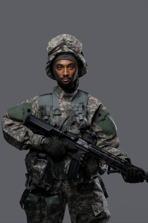Foto de Soldado de piel oscura con uniforme y casco de la OTAN posa con una expresión seria sobre un fondo gris liso - Imagen libre de derechos