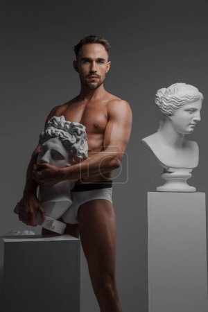 Téléchargez les photos : Un modèle masculin séduisant vêtu uniquement de sous-vêtements, posant avec une sculpture de buste grecque ancienne pressée contre son corps, sur un fond gris - en image libre de droit