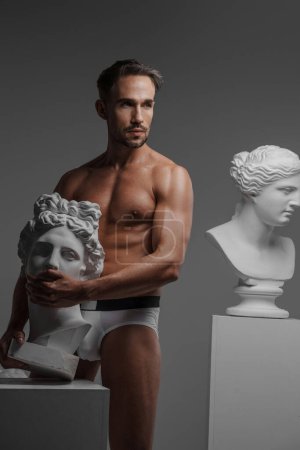 Téléchargez les photos : Un modèle masculin séduisant vêtu uniquement de sous-vêtements, posant avec une sculpture de buste grecque ancienne pressée contre son corps, sur un fond gris - en image libre de droit