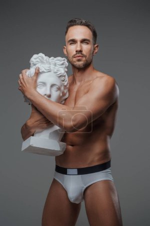 Photo for A charismatic male model with a sculpted physique poses alongside a bust of an ancient Greek sculpture - Royalty Free Image