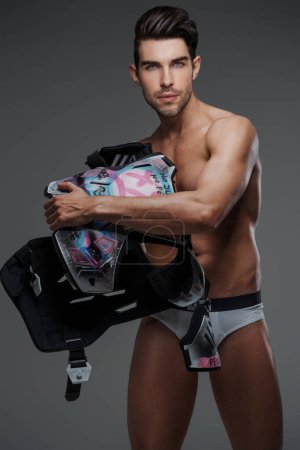 Téléchargez les photos : Brunette male model posing in underwear, holding a protective hockey gear painted in a street grunge style, against a gray background - en image libre de droit