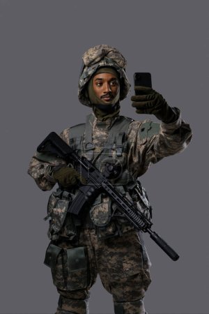 Téléchargez les photos : Young military man in uniform and helmet captures a selfie or video call with a smartphone, displaying the technology skills and adaptability of soldiers in uniform - en image libre de droit