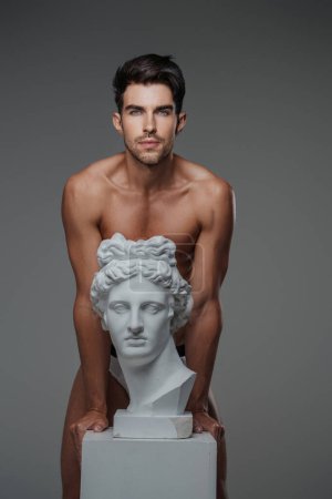 Charismatic brunette male model with a bare torso and underwear, leaning against a bust of ancient Greek sculpture, against a gray background