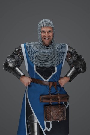 Photo for Intimidating royal guard dressed in medieval armor and blue surcoat, with chain mail coif, standing with hands on hips and a mocking expression, on a gray background - Royalty Free Image