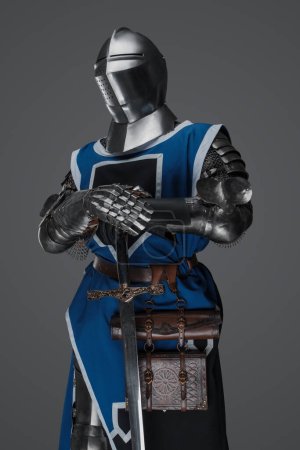Foto de Un soldado medieval vestido con un abrigo azul y una armadura sosteniendo una espada con una pose fija, similar a una estatua, sobre un fondo gris - Imagen libre de derechos