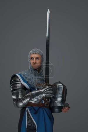 Téléchargez les photos : Fier chevalier tient son épée et son casque, revêtu d'un survêtement bleu, d'une coiffe en chaine et d'une armure médiévale, sur fond gris - en image libre de droit