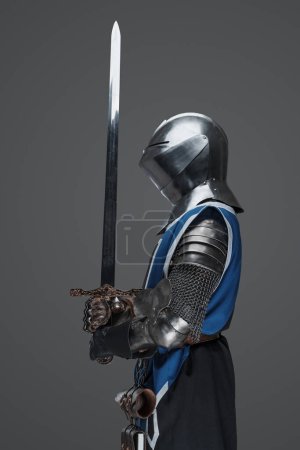 Ein mittelalterlicher Soldat in blauem Mantel und Rüstung mit einem Schwert in ruhiger, statuenartiger Pose vor grauem Hintergrund