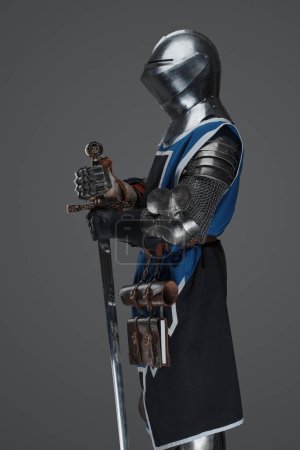 A medieval soldier garbed in blue surcoat and armor holding a sword with a still, statue-like pose, against a gray background