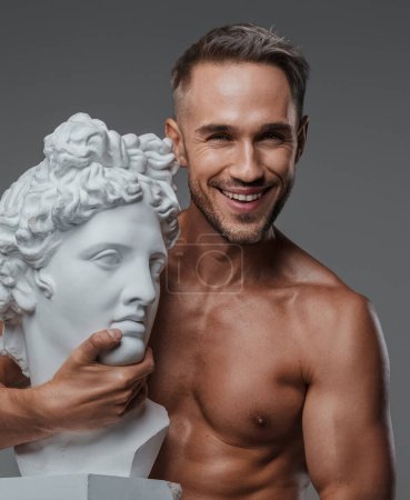 Téléchargez les photos : Un charmant modèle masculin avec un beau physique et torse nu, souriant doucement alors qu'il tient un buste d'une sculpture grecque antique sur sa poitrine, sur un fond gris - en image libre de droit