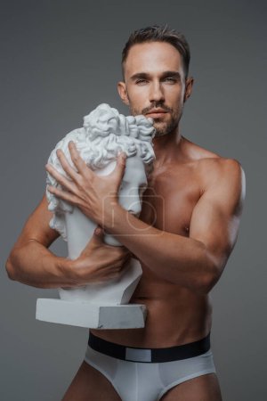 Foto de Un modelo masculino encantador, vestido solo en ropa interior, abraza un busto de una escultura griega, exudando encanto y encanto, con un fondo gris - Imagen libre de derechos