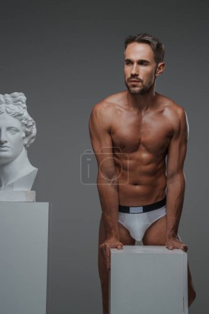 Photo for A stunning male model with a chiseled torso and in underwear striking a pose next to a bust of an ancient Greek sculpture - Royalty Free Image