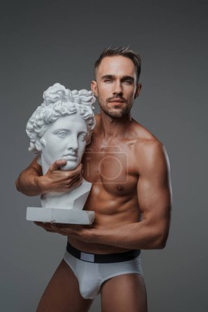 Téléchargez les photos : Un modèle masculin enchanteur, vêtu uniquement de sous-vêtements, embrasse un buste d'une sculpture grecque, respirant charme et allure, avec une toile de fond grise - en image libre de droit
