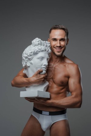 Photo for A charming male model, wearing nothing but underwear, showcases a warm smile while cradling a bust of a Greek sculpture, with a gray backdrop - Royalty Free Image