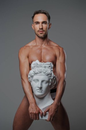Foto de Un seductor modelo masculino vestido solo con ropa interior, posando con una escultura del busto griego antiguo presionado contra su cuerpo, sobre un fondo gris - Imagen libre de derechos