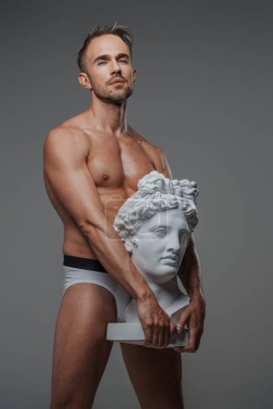Téléchargez les photos : Un modèle masculin charismatique avec un physique sculpté pose aux côtés d'un buste d'une sculpture grecque antique - en image libre de droit