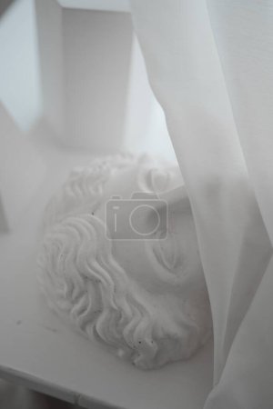 Téléchargez les photos : Photographie légèrement éclairée représentant un buste de gypse grec recouvert d'un voile translucide, posée sur une table - en image libre de droit