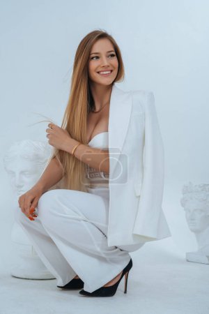 Téléchargez les photos : Portrait glamour d'une beauté statuesque en costume blanc et soutien-gorge séduisant, frappant une pose à côté d'un buste grec classique dans une séance photo de haute couture - en image libre de droit