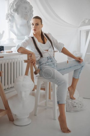 Téléchargez les photos : Sculptrice talentueuse vêtue d'une chemise blanche déboutonnée et d'un jean à bretelles, tenant un marteau et posant sur une chaise dans son atelier de sculpteur entouré de sculptures - en image libre de droit