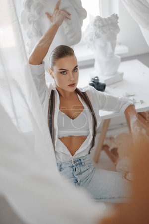 Photo for A portrait of a beautiful sculptor girl dressed in a white unbuttoned shirt and jeans with suspenders, holding a hammer, posing sitting on a chair in her sculptors workshop surrounded by sculptures - Royalty Free Image