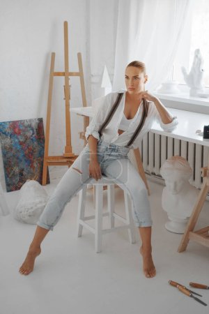 Foto de Una atractiva escultora vestida con una camisa blanca suelta y jeans con tirantes, sentada en un taburete en su estudio de escultores - Imagen libre de derechos