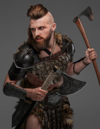 Téléchargez les photos : Un homme viking imposant et robuste avec une barbe touffue, vêtu de fourrure animale et une armure légère, tenant deux haches sur un fond gris neutre - en image libre de droit
