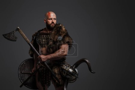 Photo for An intimidating, bald, bearded Viking donning fur and lightweight armor, keeping a helmet fastened to his belt, gripping a menacing axe and shield on a gray backdrop - Royalty Free Image