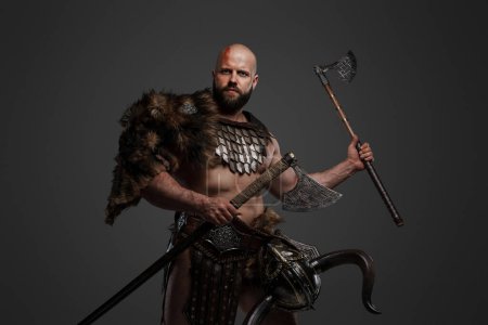 Photo for A strong and intimidating Viking man in a beard and bald head, dressed in animal fur and light armor, with a helmet hanging from his belt, brandishing a huge axe against a gray background - Royalty Free Image