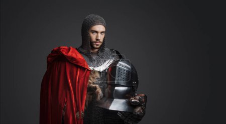 Photo for Regal portrait of a young king in armor with steel plates, holding a steel helmet and wearing a red cloak, posing on a neutral background - Royalty Free Image