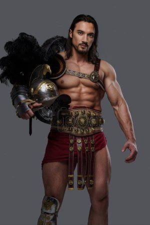 Téléchargez les photos : Gladiateur puissant avec une barbe élégante porte une armure complexe légère et pose serrant un casque à plumes sur un fond gris - en image libre de droit