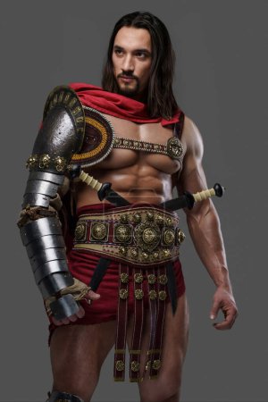Foto de Potente gladiador con una elegante barba lleva una armadura ligera e intrincada y posa agarrando un casco emplumado sobre un fondo gris - Imagen libre de derechos