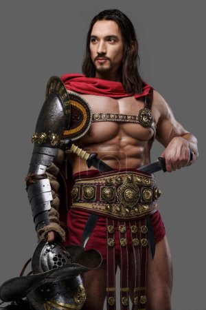 Foto de Gladiador muscular con una barba elegante y el pelo que fluye dons armadura ligera intrincada, sostiene un casco emplumado sobre un fondo gris - Imagen libre de derechos
