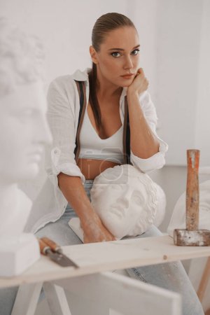 Téléchargez les photos : Sculptrice talentueuse dans une chemise blanche et un jean, tenant un buste grec antique dans son atelier, entourée d'outils. Un mélange parfait d'artisanat et d'art - en image libre de droit