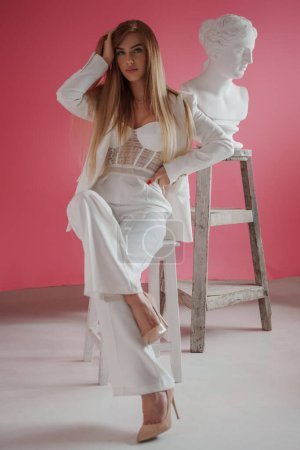 Foto de Modelo femenina de moda y seductora en un traje blanco y top de sujetador sexy posando en un estudio fotográfico con un busto griego antiguo, sobre un fondo rosa - Imagen libre de derechos