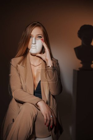 Photo for Beautiful woman in an elegant beige suit, partially hiding her face with a plaster ancient Greek mask against a warm illuminated background, with the silhouette of an ancient Greek bust visible - Royalty Free Image