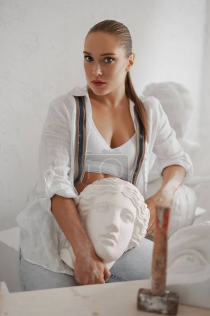 Foto de Talentosa escultora con camisa blanca y jeans, sosteniendo un antiguo busto griego en su taller, rodeada de herramientas. Una mezcla perfecta de artesanía y arte - Imagen libre de derechos