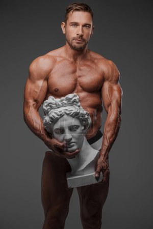 Foto de Un guapo modelo masculino en ropa interior mostrando una pose cautivadora con un busto griego antiguo sobre un fondo gris - Imagen libre de derechos