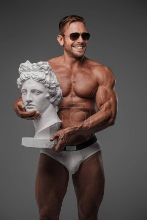 Photo for A charismatic male model in sunglasses and underwear showcasing a wide smile while holding an ancient Greek bust on a gray background - Royalty Free Image