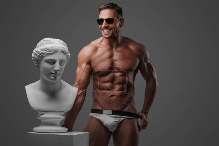 Foto de Sonriente modelo masculino musculoso atractivo usando gafas de sol y ropa interior se coloca con confianza al lado del busto griego antiguo sobre un fondo gris - Imagen libre de derechos
